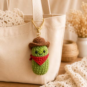 May include: A crochet pickle keyring featuring a green textured body, a brown cowboy hat, and a red bandana. The plush figure has two small black eyes and a stitched smile, attached to a gold-toned metal lobster clasp hanging from a canvas bag.