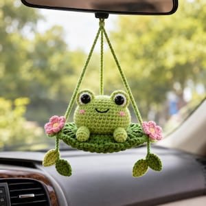 May include: A crochet car hanging ornament featuring a light green frog sitting on a dark green lily pad. The lily pad is suspended by green yarn strings and decorated with two pink flowers with yellow centers and two green leaves on each side.