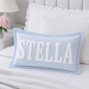 May include: A rectangular light blue throw pillow featuring the name STELLA in large, white, textured, fuzzy block letters centered on the front. The pillow has a piped edge border and a soft, smooth fabric finish across the entire surface.