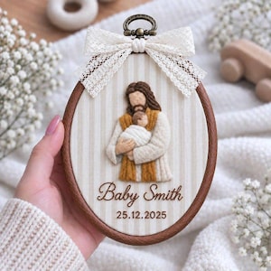 Puede incluir: Un bastidor de bordado ovalado con un fondo de tela de rayas verticales beige. Una figura de lana fieltrada de una persona sosteniendo a un bebé envuelto está centrada. Debajo, el texto dice Baby Smith y 25.12.2025. Un lazo de encaje blanco está sujeto.