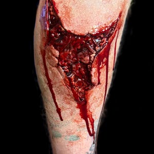 May include: A realistic prosthetic special effects makeup application depicting a deep, jagged laceration on skin. The wound features exposed textured tissue in shades of dark red and brown, with bright red liquid blood dripping down the surface.