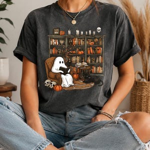 Op de afbeelding: Een antracietgrijs, mineraal gewassen t-shirt met een grafische print van een witte geest die in een bruine fauteuil zit te lezen, omringd door een boekenplank vol boeken, pompoenen, kaarsen, een schedel en een zwarte kat op de vloer.