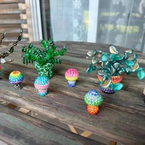 May include: A collection of three beaded plant sculptures in pots and five small mushroom-shaped ornaments. The plants feature green and dark metallic beads, while the pots and mushrooms are covered in multicolored, vibrant, sparkling rhinestones.