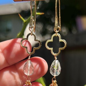 May include: A pair of gold-toned earrings featuring a four-lobed clover-shaped metal frame, a clear faceted teardrop-shaped gemstone, and a cluster of small pink beads at the base, all suspended from long gold-toned ear wires.