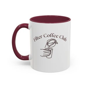 May include: A white ceramic mug with a dark maroon handle and interior rim. The front features a black line art illustration of a hand pouring liquid into a cup, accompanied by the text Filter Coffee Club in a simple, dark serif font.