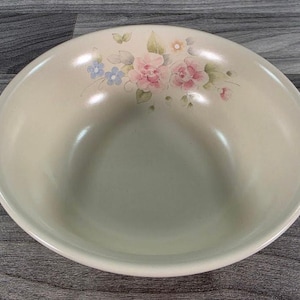 May include: A round, cream-colored ceramic bowl featuring a delicate floral pattern near the rim. The design includes soft pink roses, small blue flowers, and green leaves painted in a watercolor style. The bowl has a smooth, glossy finish.