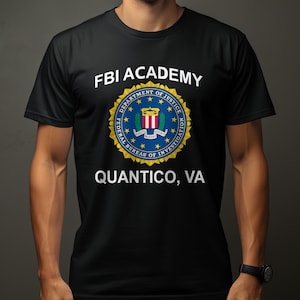 Op de afbeelding: Een zwart t-shirt met korte mouwen voorzien van de tekst FBI ACADEMY in het wit boven een cirkelvormig blauw, goud en wit zegel van de Federal Bureau of Investigation, met de tekst QUANTICO, VA in het wit onder het zegel op de borst.