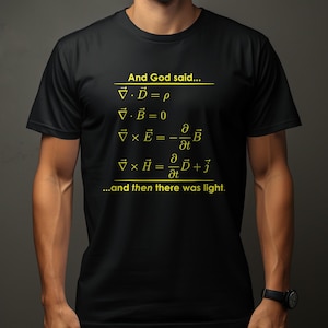 Op de afbeelding: Een zwart T-shirt met korte mouwen, voorzien van gele tekst en wiskundige vergelijkingen. De opdruk toont de tekst "And God said..." gevolgd door de vergelijkingen van Maxwell en de slotzin "...and then there was light." in het geel.
