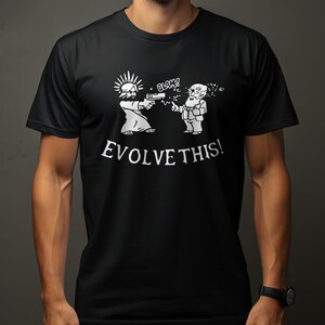 Puede incluir: Una camiseta negra de manga corta con un estampado gráfico blanco de una figura con halo apuntando con una pistola a un hombre barbudo. El texto "BLAM!" aparece sobre el arma y las palabras "EVOLVE THIS!" están impresas en blanco debajo.