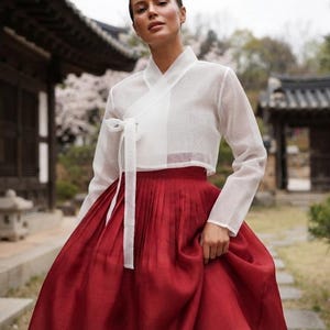以下が含まれることがあります： 伝統的な韓国の韓服（ハンボク）で、白い長袖の半透明なラップスタイルのジャケットにフロントタイの留め具が付いており、ハイウエストで床まで届く長さの深い赤色の無地のプリーツスカートと組み合わされています。
