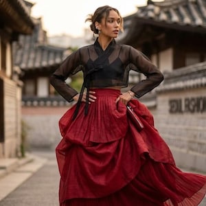 Puede incluir: Una blusa cruzada negra transparente de manga larga con cierre de lazo sobre una camiseta negra, combinada con una falda plisada de color rojo oscuro, voluminosa y hasta el suelo. La falda tiene varias capas de tela fluida.