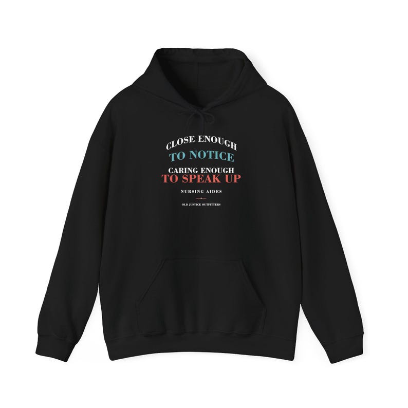 Black pullover hoodie, front view, printed CLOSE ENOUGH TO NOTICE, CARING ENOUGH TO SPEAK UP, NURSING AIDES, OLD JUSTICE OUTFITTERS.