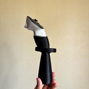 May include: A white and black gaming controller handle attachment featuring a textured black cylindrical grip with a circular ring guard, securely holding a white and black handheld motion controller with a small black joystick at the top.