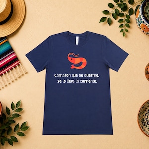 May include: A navy blue short-sleeve t-shirt featuring a graphic of an orange shrimp and the white text "Camarón que se duerme, se lo lleva la corriente." The shirt has a crew neckline and a standard fit, displaying a clear, centered design.