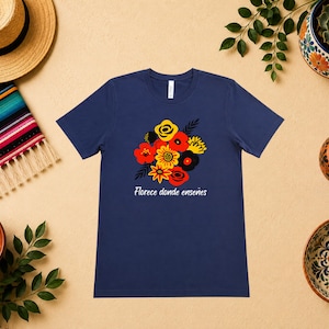 May include: A navy blue short-sleeve t-shirt featuring a central graphic of a stylized floral bouquet in red, yellow, and black. Below the flowers, the white text reads "Florece donde enseñes" in a cursive script font on the front of the shirt.