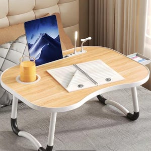 May include: A portable light wood grain laptop desk with white curved legs and black non-slip feet. The desk features a built-in cup holder, a tablet slot, a small side drawer, and a white LED lamp. A notebook and a glass of orange juice are on top.