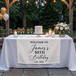 May include: A white table runner featuring the black printed text "WELCOME TO James's 50TH Birthday EST. 1976" draped over a table with a cake, candles, champagne glasses, and a bouquet of white roses under a wooden pergola with string lights.
