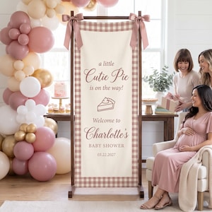 May include: A vertical rectangular banner with a dusty rose and white gingham border, featuring the text 'a little Cutie Pie is on the way!', a pie slice icon, 'Welcome to Charlotte's BABY SHOWER', and '03.22.2027', suspended from a wooden frame with satin bows.