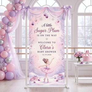 May include: A vertical baby shower banner featuring a ballerina in a pink tutu, surrounded by ornaments, snowflakes, and gifts. The text reads A little Sugar Plum IS ON THE WAY WELCOME TO Clara's BABY SHOWER 12.19.2026. It is framed by a balloon arch.
