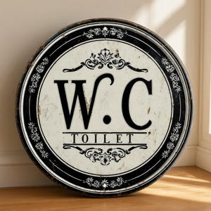 May include: A circular sign with a distressed off-white center and a black border. The sign features the large black text W.C. above the word TOILET in a smaller font. Decorative black scrollwork and floral patterns frame the text.