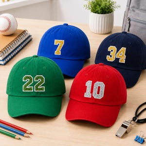 Puede incluir: Cuatro gorras de béisbol en verde, azul, rojo y azul marino. La gorra verde presenta el número 22 en purpurina verde, la azul el número 7 en purpurina dorada, la roja el número 10 en purpurina plateada y la azul marino el 34 en dorado.
