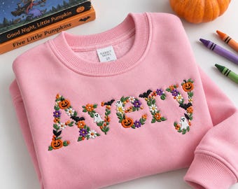 May include: A light pink long-sleeve sweatshirt featuring the name AVERY embroidered on the front in a floral and Halloween-themed font. The letters are decorated with small orange pumpkins, white ghosts, black bats, and colorful flowers.