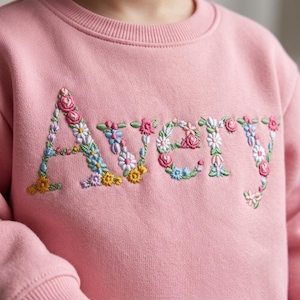 Puede incluir: Una sudadera de cuello redondo en color rosa claro con el nombre AVERY bordado en el pecho. Las letras están formadas por un colorido patrón floral compuesto por pequeñas flores rosas, azules, amarillas y blancas con hojas y tallos verdes.