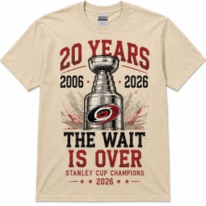 May include: A beige short-sleeve t-shirt featuring a graphic of a hockey trophy with the text 20 YEARS 2006 2026 THE WAIT IS OVER STANLEY CUP CHAMPIONS 2026. The design includes a red and black circular logo on the trophy and red star accents.