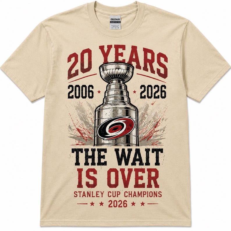 May include: A beige short-sleeve t-shirt featuring a graphic of a hockey trophy with the text 20 YEARS 2006 2026 THE WAIT IS OVER STANLEY CUP CHAMPIONS 2026. The design includes a red and black circular logo on the trophy and red star accents.