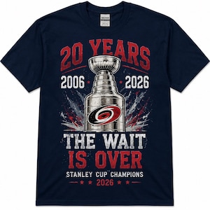 May include: A navy blue short-sleeve t-shirt featuring a graphic of a silver trophy cup with a red and black logo. Text reads 20 YEARS, 2006, 2026, THE WAIT IS OVER, STANLEY CUP CHAMPIONS, and 2026, accented with red stars and a distressed design.