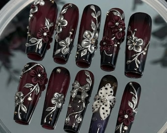 May include: A set of ten long, coffin-shaped press-on nails featuring a deep burgundy and black gradient base. The nails are decorated with three-dimensional silver floral embellishments, metallic vines, small clear rhinestones, and a white bow.