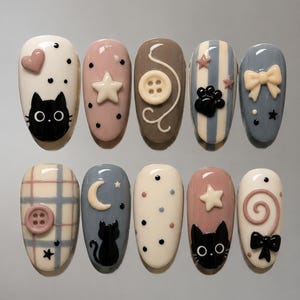 May include: A set of ten press-on nails featuring various designs including black cat silhouettes, cream-colored stars, buttons, bows, a crescent moon, and a spiral. The color palette includes cream, dusty pink, brown, blue, and grey with polka dot patterns.