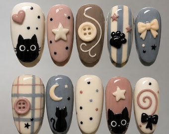 May include: A set of ten press-on nails featuring various designs including black cat silhouettes, cream-colored stars, buttons, bows, a crescent moon, and a spiral. The color palette includes cream, dusty pink, brown, blue, and grey with polka dot patterns.