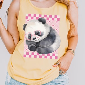 Puede incluir: Un top sin mangas amarillo con un gráfico de un oso panda con gafas de sol de corazón rosa. El panda está durmiendo sobre un fondo a cuadros rosa y blanco con corazones rosas.
