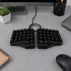May include: A black split ergonomic mechanical keyboard with two separate halves connected by a black cable. The keys are black with white lettering in a QWERTY layout. The device features a contoured shape and is positioned on a flat surface.