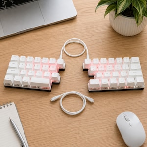 May include: A split ergonomic mechanical keyboard with white keycaps and pink backlighting. The two halves are connected by a white coiled cable, with a second white cable positioned between them. The keyboard features a standard QWERTY layout.