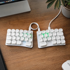 May include: A split ergonomic keyboard featuring two separate white rectangular sections with rounded white keys. Each key displays a black letter or command label. The device is connected by a white cable and emits a soft green light from beneath the keys.