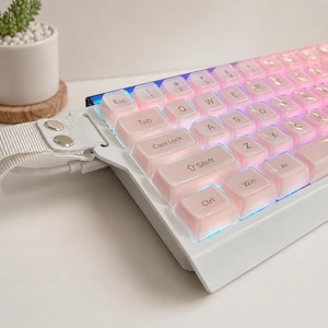 May include: A white mechanical keyboard featuring translucent light pink keycaps with black lettering and integrated RGB lighting. The keyboard includes a white fabric strap attached to the left side with two silver metal rivets and a white cable.