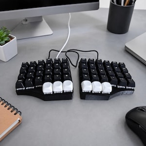 May include: A split ergonomic mechanical keyboard featuring two separate black sections with black keycaps and three white thumb keys on each side. The keyboard is connected by a black cable and includes white lettering on the keys for standard QWERTY layout.