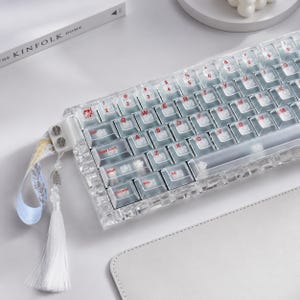 May include: A mechanical keyboard with a transparent acrylic case and clear keycaps featuring red lettering. The keyboard includes a decorative white tassel and a patterned fabric strap attached to the left side with two silver-colored metal screws.