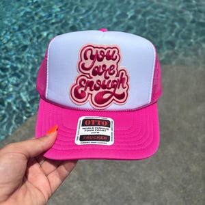 Pode incluir: Um boné trucker rosa choque e branco com um painel frontal de espuma branca e um emblema bordado em rosa que diz "You are Enough" em letra cursiva. O boné inclui uma parte traseira de rede rosa, uma aba rosa e um autocolante na aba.