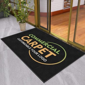 May include: A rectangular black floor mat featuring the text COMMERCIAL CARPET in large tan letters and CUSTOMIZABLE YOUR LOGO in smaller white letters, all enclosed within a circular graphic outline that transitions from green to tan.