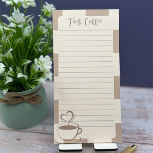 May include: A rectangular notepad with a cream background and brown border accents. The top features the text First Coffee in a script font. Below, horizontal lines fill the page, and a brown line drawing of a coffee cup with steam in a heart shape sits at the bottom left corner.