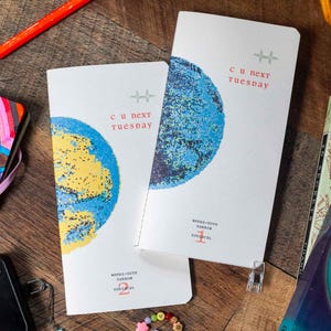 May include: Two white rectangular notebooks featuring a graphic of a blue and yellow planet. The text "c u next tuesday" is printed in red, with "weeks-note narrow" and "zodiacal" below. One notebook is marked with a red 1 and the other with a 2.