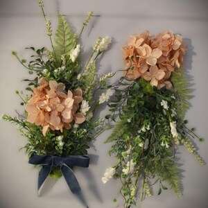 May include: Two floral arrangements featuring peach-colored hydrangea blossoms, green fern fronds, and small white flowers. The left arrangement is accented with a dark blue velvet ribbon tied in a bow at the base of the stems and foliage.