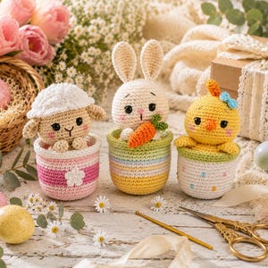 May include: Three crocheted amigurumi figures in small baskets: a white lamb in a pink basket, a cream rabbit holding an orange carrot in a pastel striped basket, and a yellow chick with a blue bow in a white basket with colorful dots.