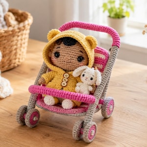 Puede incluir: Una muñeca de ganchillo con cabello oscuro que lleva una chaqueta amarilla con capucha con orejas de oso y tres botones pequeños, sentada en un cochecito de juguete de ganchillo rosa y gris. La muñeca sostiene un pequeño conejo blanco.