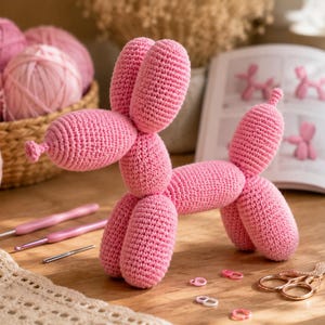 May include: A pink crocheted balloon dog figure made with yarn, featuring a rounded snout, tall ears, and a cylindrical body. The figure is crafted with a tight stitch pattern, mimicking the appearance of a twisted balloon animal sculpture.