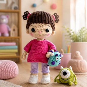 May include: A crocheted doll with brown hair in pigtails, wearing a bright pink shirt, light purple pants, and white shoes. The doll holds a small blue monster-themed bag and stands next to a small green one-eyed monster crocheted figure.