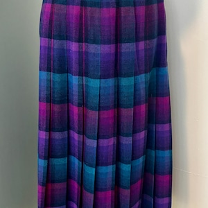 May include: A midi-length pleated skirt featuring a plaid pattern in shades of magenta, purple, and teal. The skirt has a fitted waistband and vertical pleats that extend from the waist to the hem, creating a structured and uniform silhouette.